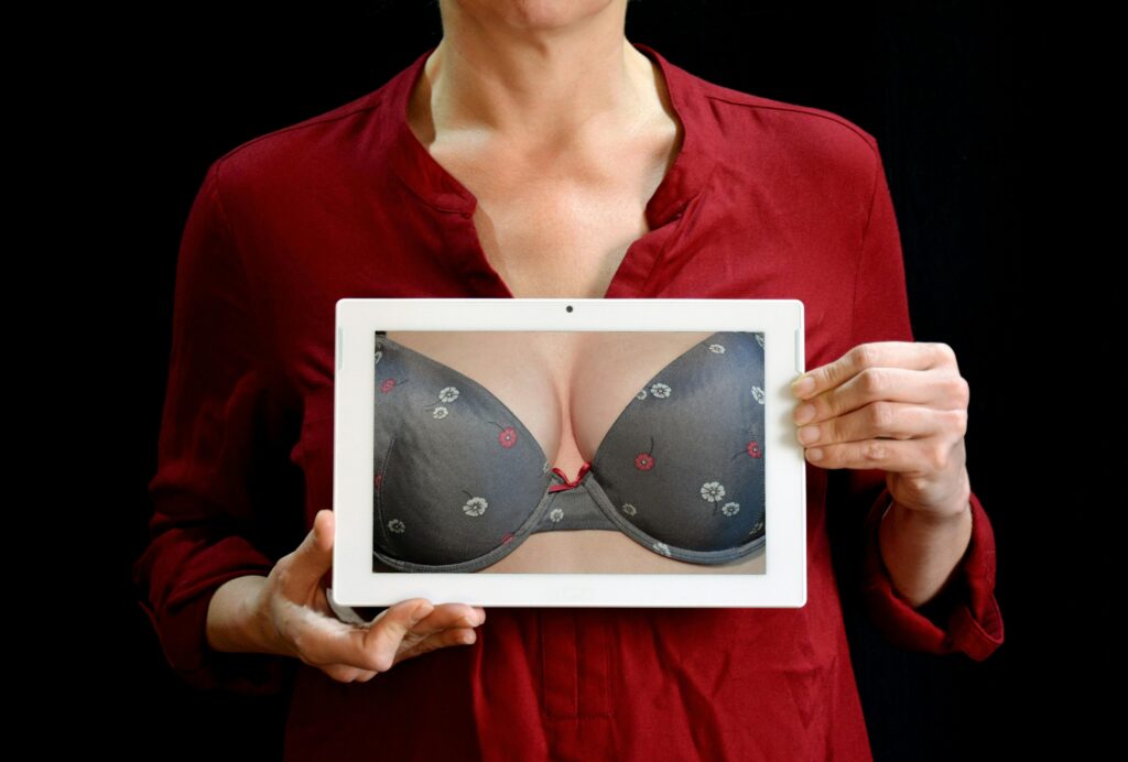 Person in a red blouse holds a white tablet showing a gray floral bra against a black background.