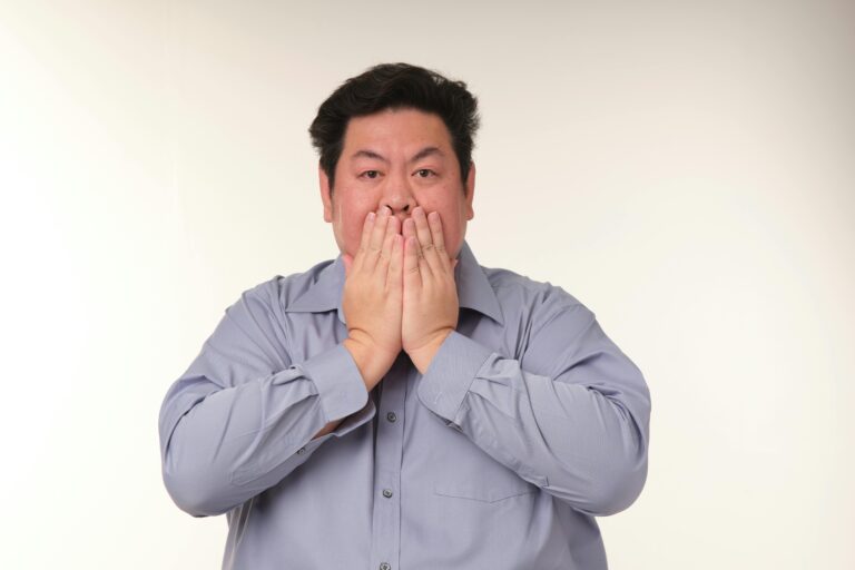 Front-facing man in a light blue shirt covers his mouth with both hands, eyes widened in surprise.
