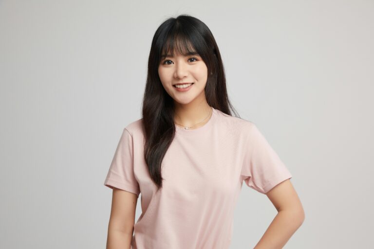 Portrait of a smiling woman with long dark hair wearing a pale pink t-shirt against a light gray backdrop.