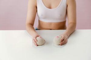 Person in a white sports bra sits at a white table, holding two knitted beige balls in front of a pink wall.