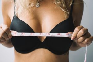 Close-up of a woman measuring her bust with a white measuring tape while wearing a black bra, necklace visible.