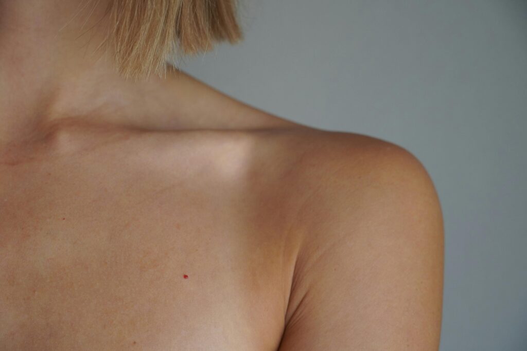 Back of a person’s shoulder and neck with short blond hair at the nape against a gray backdrop.