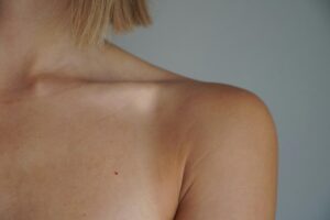 Back of a person’s shoulder and neck with short blond hair at the nape against a gray backdrop.