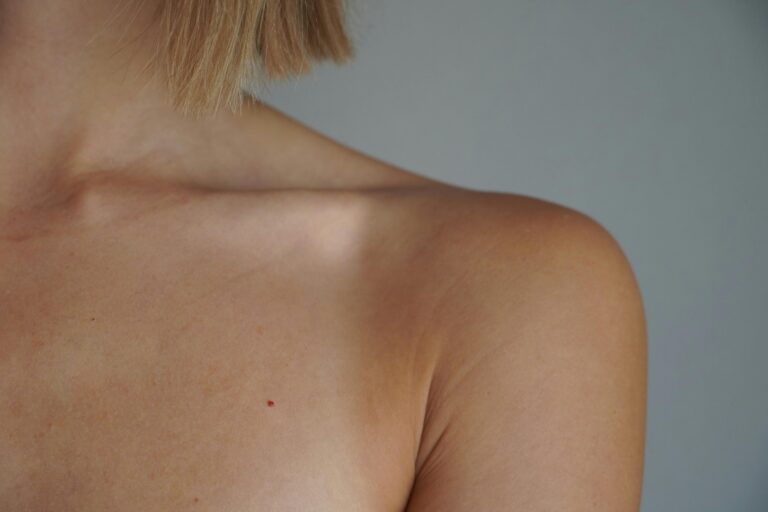 Back of a person’s shoulder and neck with short blond hair at the nape against a gray backdrop.