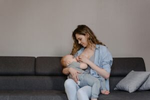 Mother seated on a dark sofa while nursing her baby, both in casual clothes in a cozy living room setting.