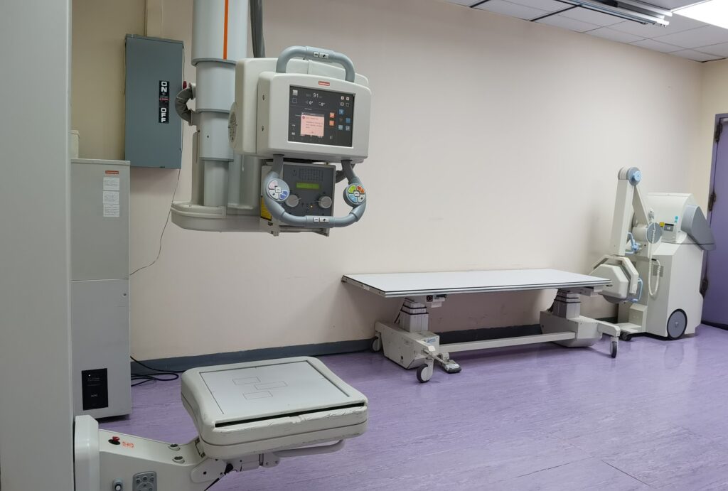 Hospital radiology room with a ceiling-mounted imaging device and control console above a patient table, used for diagnostic X-ray imaging.