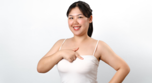 Smiling woman in a white tank top points to her chest against a light background.