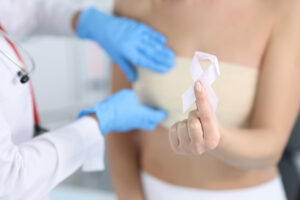 Hand in foreground holding a pink breast cancer awareness ribbon, with a clinician wearing blue gloves in the background.