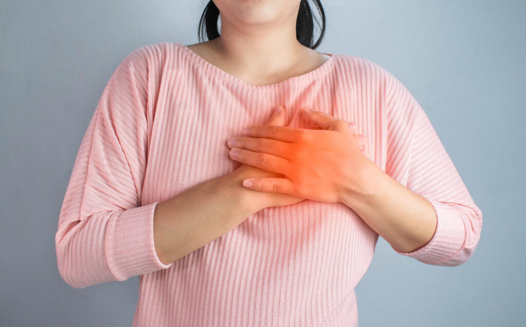 Person in a pink sweater places both hands on their chest, with a red glow indicating chest pain or discomfort on the chest area.