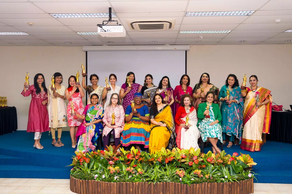 Group of women in colorful traditional dresses posing on a stage, each holding a trophy after an award ceremony, with a flower bed in front.