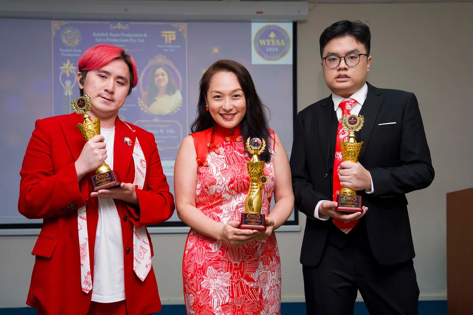 Three award winners pose with gold trophies at an indoor ceremony; left man in red suit, center woman in red dress, right man in black suit.