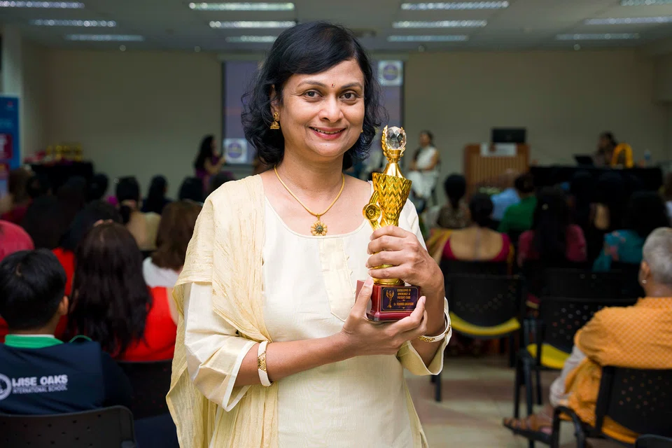 Smiling woman in a cream outfit holds a gold trophy at an award ceremony with a seated audience in the background.