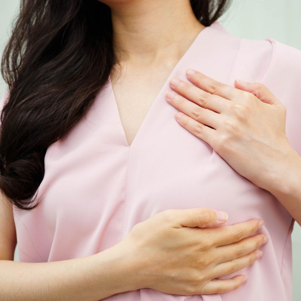 Close-up of a woman in a pink blouse performing a breast self-exam, with one hand on her chest and the other on her breast area.