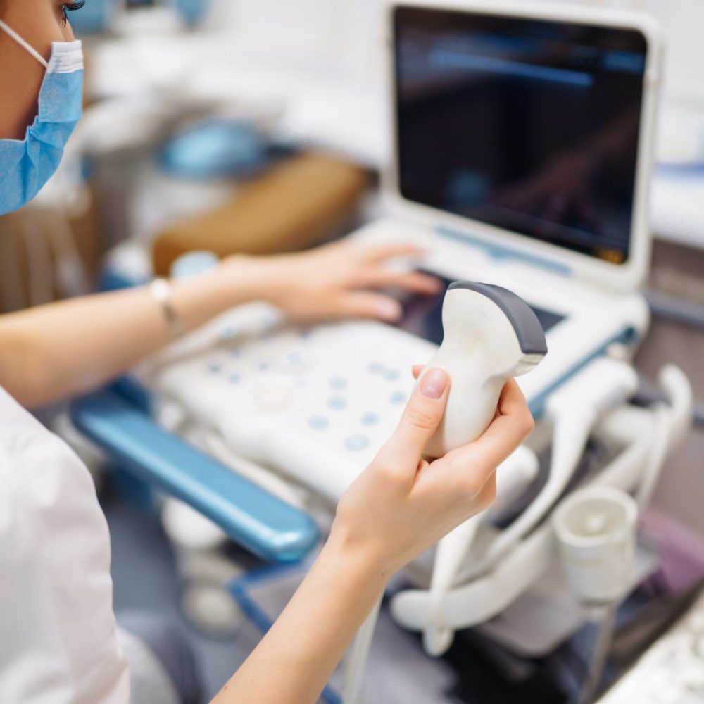 Medical professional wearing a mask uses an ultrasound probe with a monitor in view in the background.